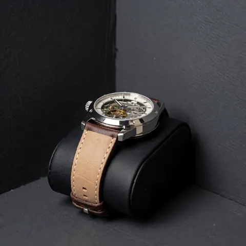 Luxury mechanical wristwatch with leather strap and visible movement through the glass, displayed on a black cushion against gray walls with soft lighting