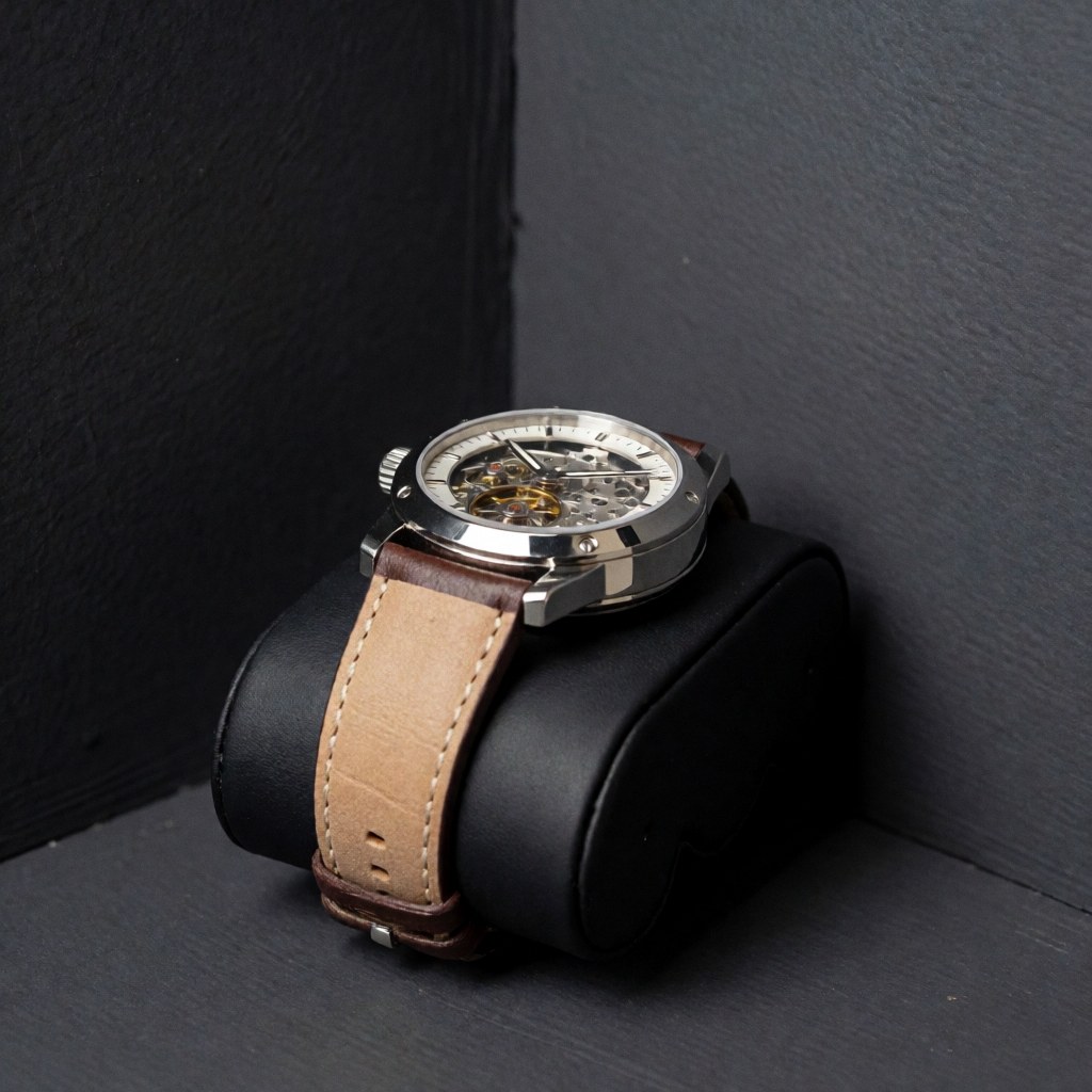 Luxury mechanical wristwatch with leather strap and visible movement through the glass, displayed on a black cushion against gray walls with soft lighting