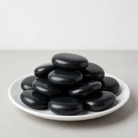 Polished black basalt stones arranged on a white ceramic plate against a neutral gray background with soft lighting
