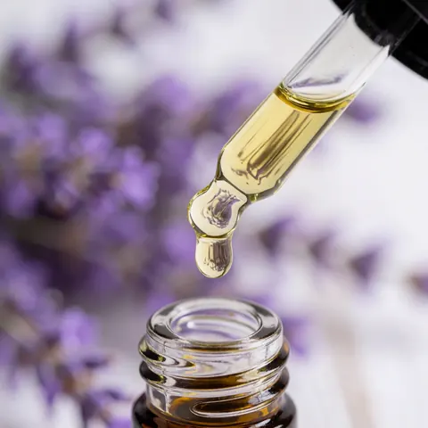 A single drop of lavender essential oil falling into a clear glass bottle with a blurred background of lavender flowers and white canvas for hot stone massage products