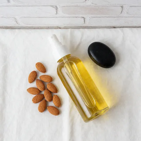 Spray bottle of almond oil, whole almonds, and a black hot stone arranged on a white cloth for spa and massage therapy