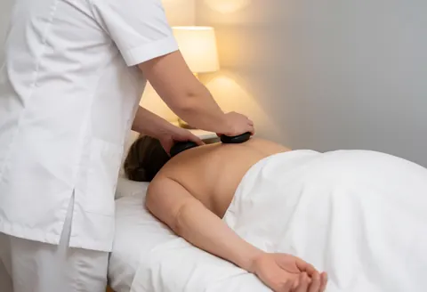 A professional masseuse in white uniform applies hot stones to a client's back on a towel-covered table under warm lighting.