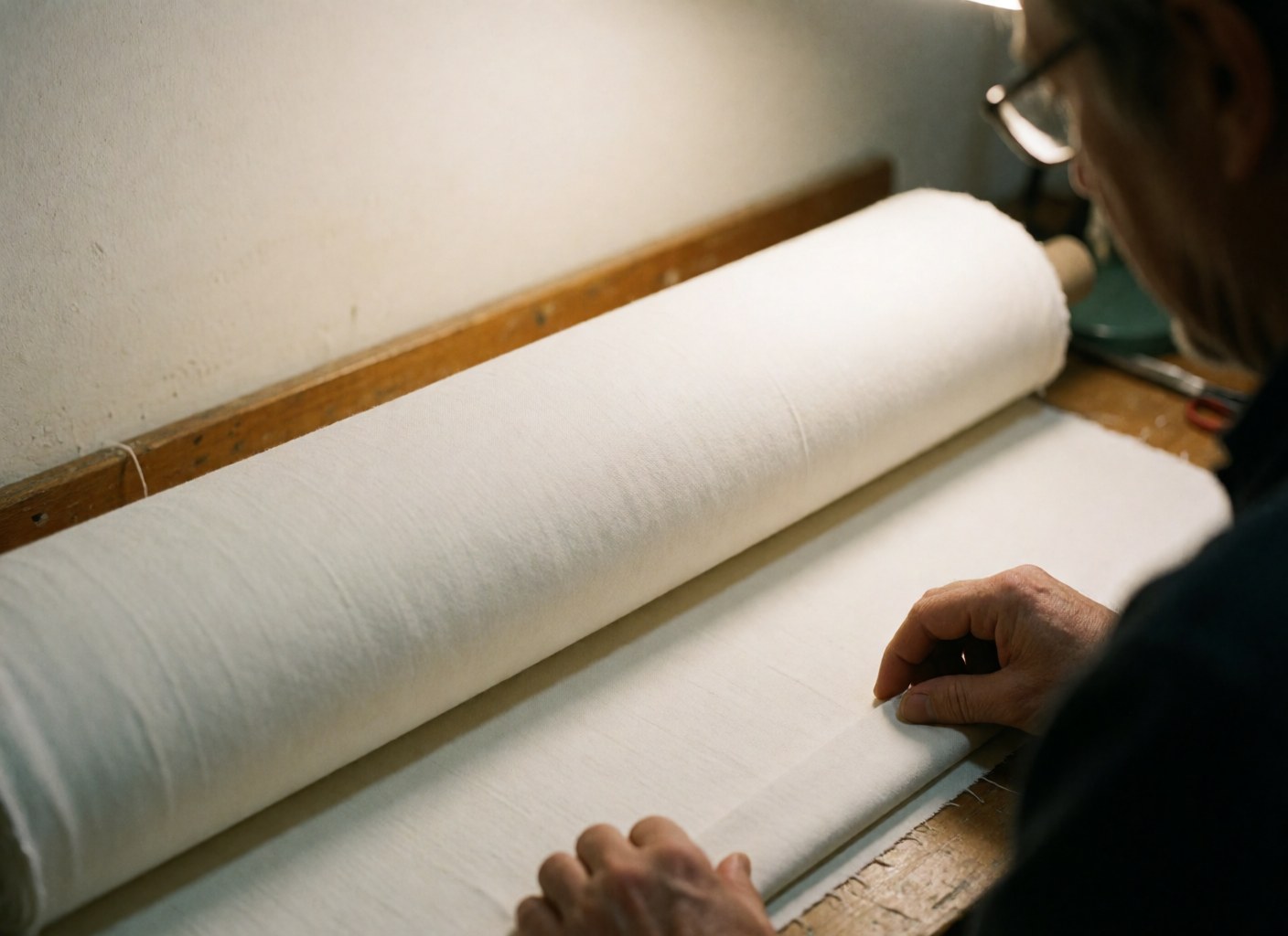 Close-up of a white fabric roll on a wooden workbench with hands working nearby, warm lighting, and white walls in a textile workshop.