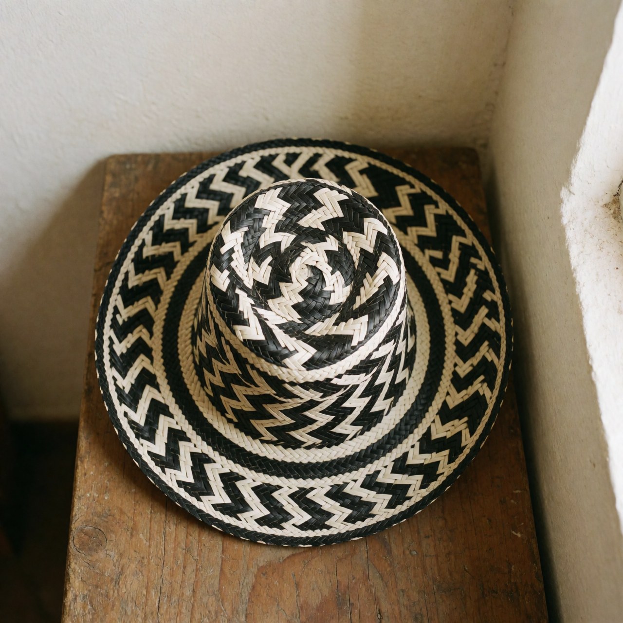 A traditional black and white woven Vueltiao hat resting on a rustic wooden bench against a white wall with natural light