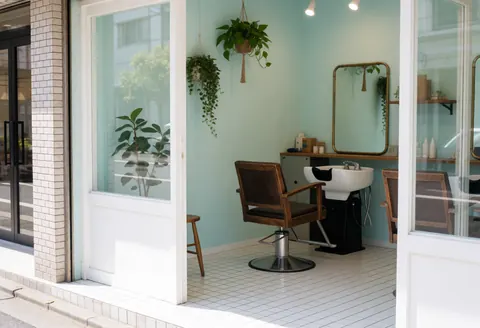 View of a hair salon interior featuring a barber chair, white sink, bronze mirror, hanging plants, turquoise walls, and white floor.