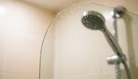 A close-up view of a curved glass shower door with water droplets and metal faucet, creating a clean and relaxing background suitable for text.