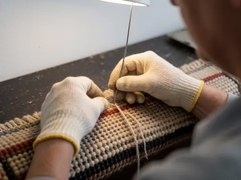 Gloved hand using a needle to stitch a carpet, highlighting wool texture and manual repair work against a dark background with warm lighting.