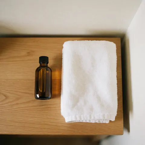 Amber glass bottle of massage oil next to a folded white towel on a light wooden table with soft natural lighting.