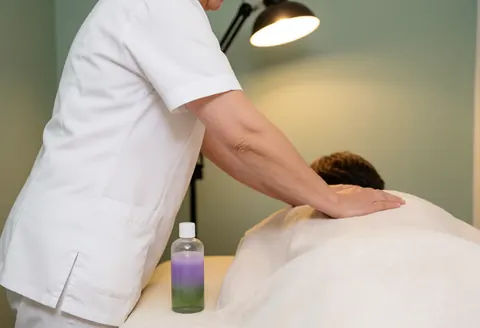 Therapist giving a Swedish massage on a table with a towel-covered client, essential oils nearby, warm lighting, and a pale green wall.