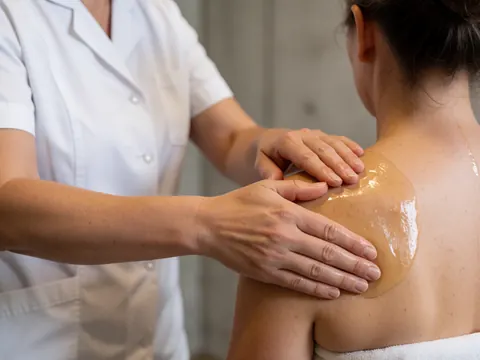 Close-up of a therapist's hands massaging a client's shoulder with oil, wearing a white uniform in a warm, relaxing environment with concrete walls.