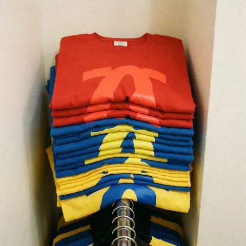 A pile of vibrant red, blue, and yellow custom screen-printed t-shirts stacked on a metal rack against a plain wall, showcasing apparel for retail sale.