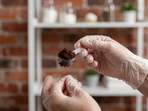 Gloved hand holding a spoon with chocolate scrub against a brick wall and white shelf background