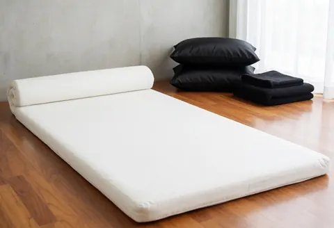 Empty Thai massage room featuring a white mattress, black pillows, polished wood floor, concrete walls, and natural light from a window.