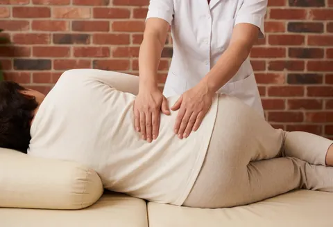 A therapist performs a Thai massage on a pregnant woman lying on a massage table in a studio with a brick wall background.