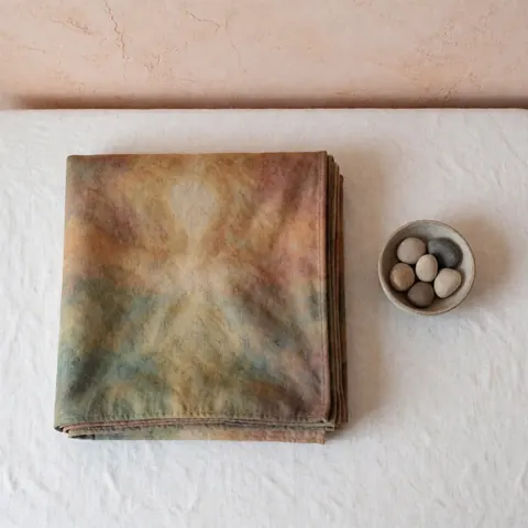 Folded Reiki blanket with subtle earthy tones placed next to a bowl of smooth stones on white fabric against a terracotta wall