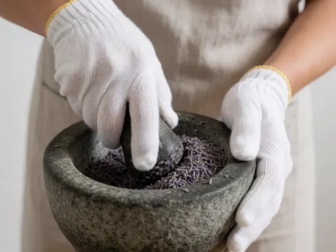 Close-up of hands using a pestle to grind dried lavender flowers inside a textured stone mortar, highlighting natural herbal preparation details