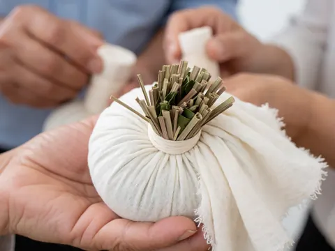 Close-up view of a Thai herbal massage compress containing dried lemongrass and kaffir lime leaves, resting on white cotton cloth with hands applying pressure in a therapeutic setting