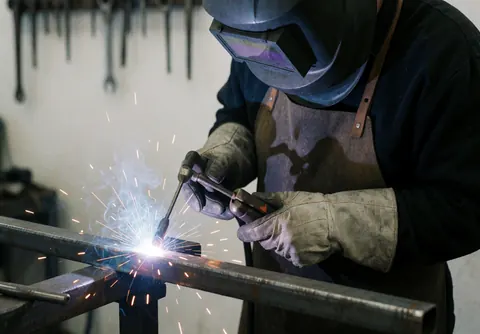 Welder wearing helmet and gloves performing arc welding on a steel bar with sparks flying and tools hanging on a white workshop wall