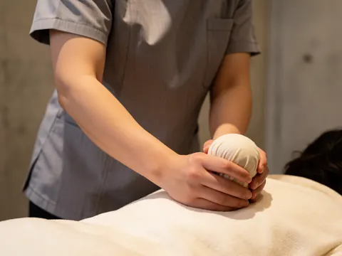 Professional therapist in gray uniform using a hot herbal compress ball on a client's back against a concrete wall with warm lighting.