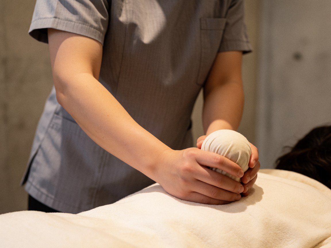 Professional therapist in gray uniform using a hot herbal compress ball on a client's back against a concrete wall with warm lighting.