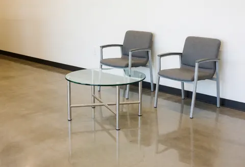 Modern waiting space featuring two gray chairs and a glass table on polished concrete floor with white walls and fluorescent lighting for copy center