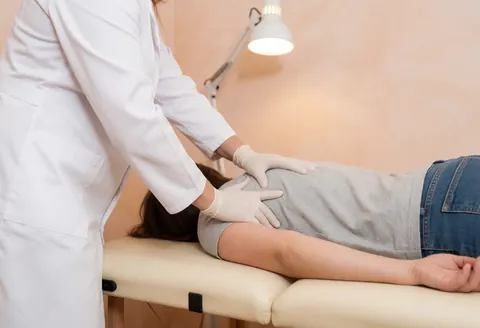 A therapist wearing gloves performs manual therapy on a patient's shoulder in a clean clinic with warm lighting and terracotta walls.
