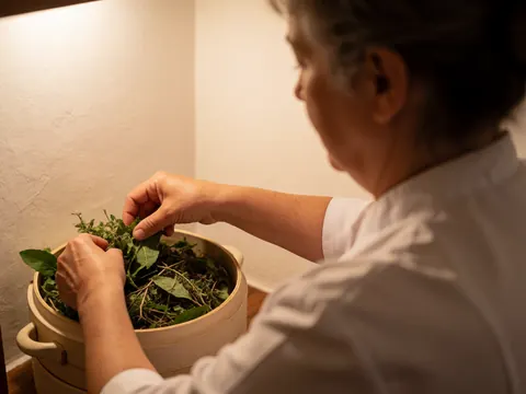 Hands placing fresh herbs into a ceramic bowl on a white surface with warm lighting for a relaxing ayurvedic therapy setting
