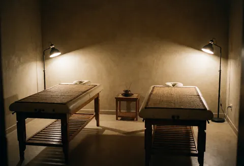 Empty ayurvedic massage room featuring two wooden treatment tables, floor lamps, and a side table with incense for a relaxing atmosphere