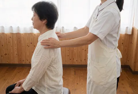 Shiatsu therapist in white uniform massages client's shoulders while seated, featuring natural light and light wood walls for a professional wellness setting