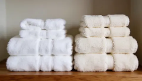 Two stacks of white and natural towels on a wooden shelf with a soft blurred background suitable for ayurvedic massage and wellness text overlays.