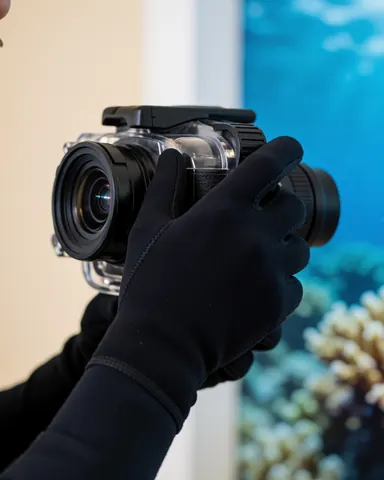 Close-up of hands wearing black gloves holding a professional waterproof camera lens, ready for underwater photography near coral reefs