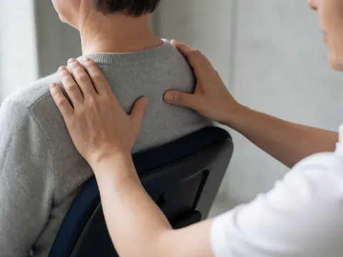 Close-up of hands applying shiatsu pressure points on a client's shoulders while seated in an ergonomic chair