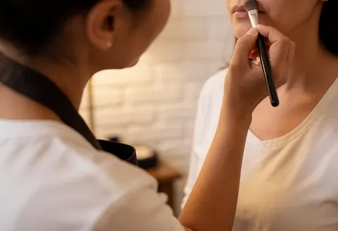 A professional makeup artist uses a brush to apply makeup to a client in a warm setting with white brick walls