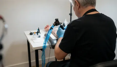 Professional tattoo artist wearing blue gloves applying ink to an arm on a white table with sterile equipment and a clean background