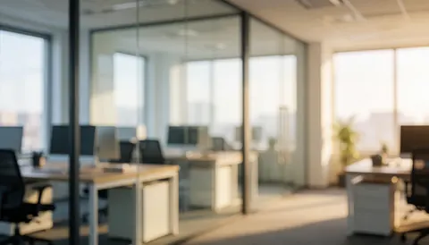 A modern office interior featuring glass partitions, work desks, chairs, and warm lighting creating a professional atmosphere.