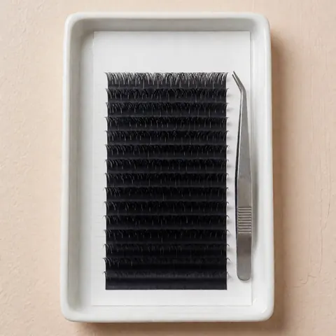 Black false eyelash extensions arranged on a white ceramic tray alongside professional tweezers, set against a soft beige background with even lighting for beauty product display