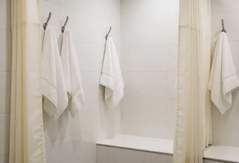 Clean shower area featuring white curtains and towels hanging on hooks, white tiled walls, and a ceramic bench for laser hair removal clients.