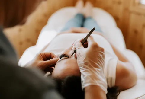 Lash technician wearing gloves applies a false eyelash to a client resting in a white treatment chair in a warm, wood-paneled salon.