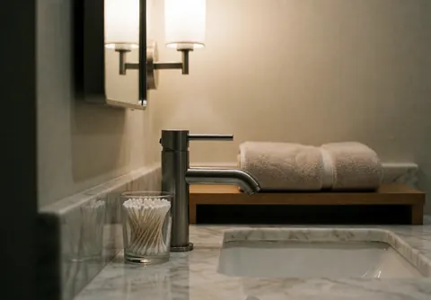 Clean modern bathroom featuring a marble sink with a stainless steel faucet, folded towels on a wooden shelf, and warm lighting.