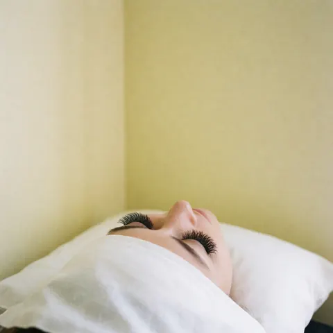 Eyes with long, voluminous false eyelashes resting on a white pillow against a light yellow wall with soft diffuse lighting