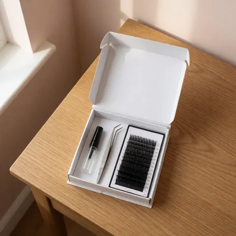 Open white box containing false eyelashes, tweezers, and glue tube on a light wooden table with natural window light