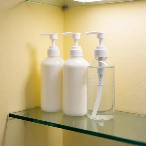 Three pump bottles, two white and one clear, sitting on a glass shelf against yellow walls with warm lighting and soft reflections
