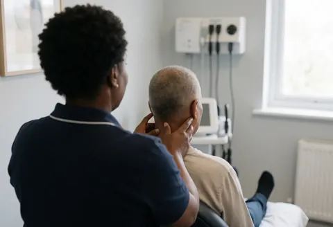 Therapist applying deep tissue massage to a client's neck while seated in a clinical setting with gray walls and medical equipment in the background.