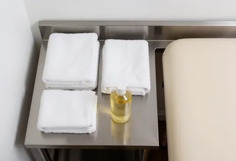 Folded white towels and a bottle of massage oil arranged on a stainless steel table next to a beige massage table in a clean professional setting