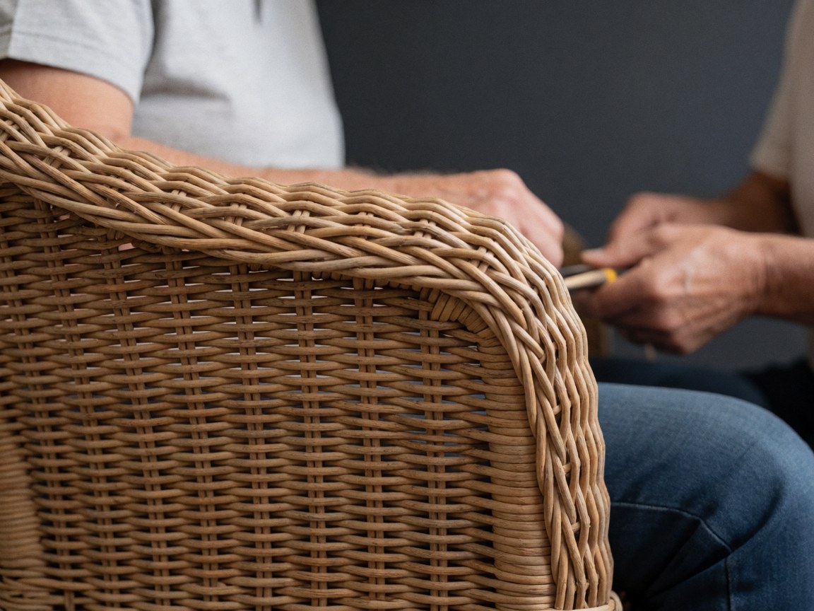 Close-up of hands using a tool to repair the arm of a wicker chair against a gray wall, highlighting natural woven texture and furniture maintenance details.