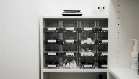 White shelving unit with clear organizers filled with eyebrow care products, a digital scale, and bottles in a clean, professional studio setting