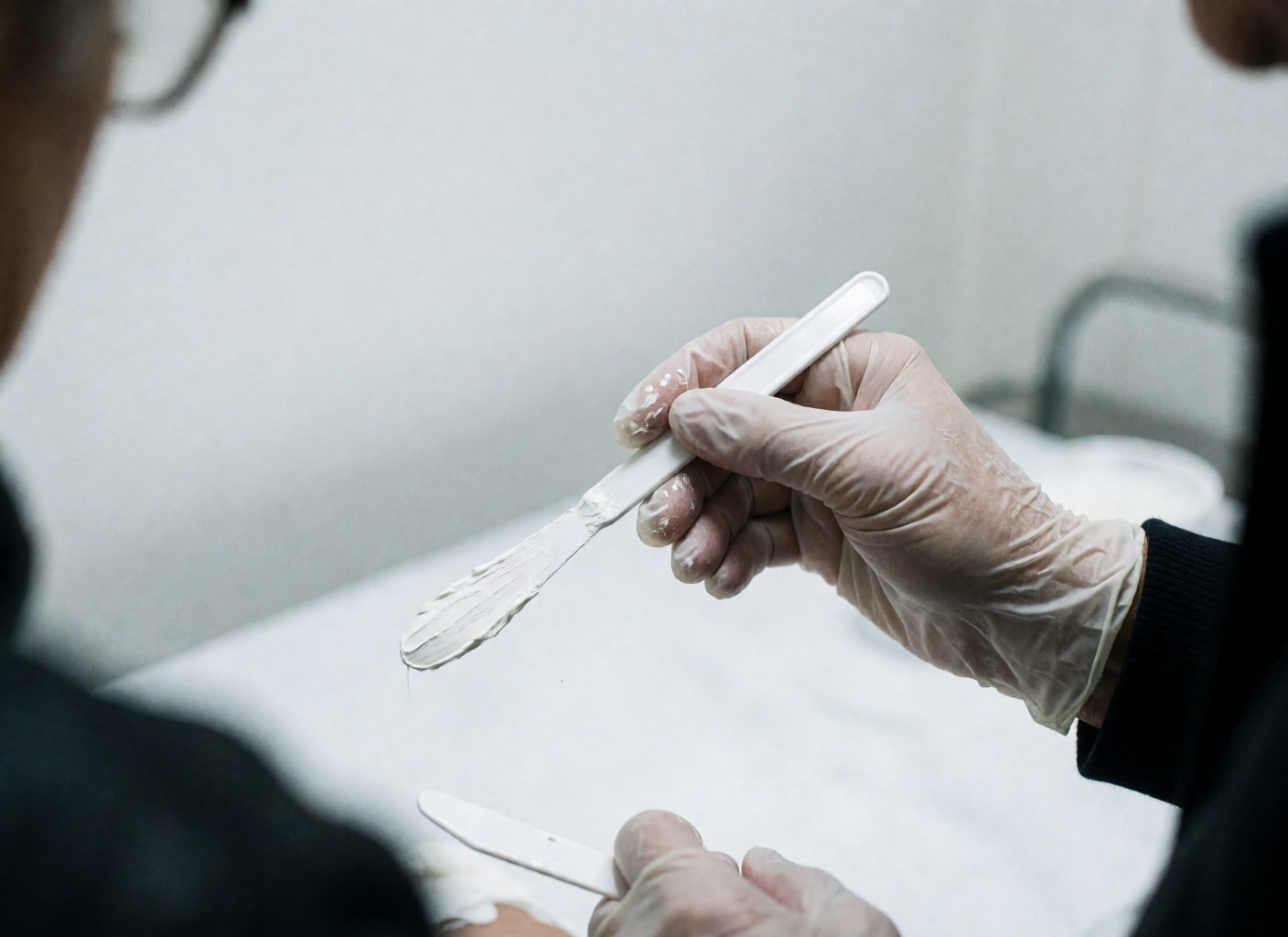 Close-up of a sterile gloved hand using a spatula to apply anesthetic cream on a clean white clinical surface under cool lighting