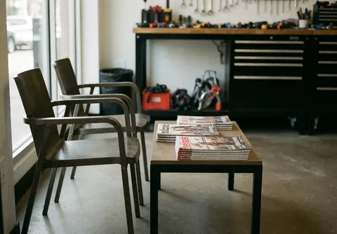 Waiting area in a bike repair shop featuring two modern metal chairs and a table with magazines, with a workbench and tools in the background.