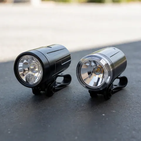 Two black bicycle headlights with clear lenses mounted on brackets on gray concrete, lit by neutral daylight, ideal for bike repair shops