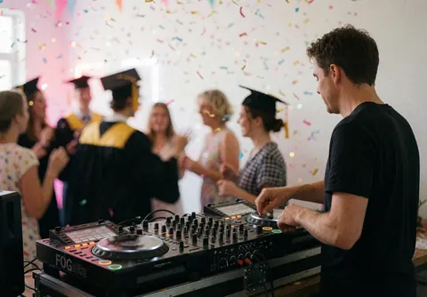 Professional DJ mixing music at a graduation party with people dancing in the background, confetti, and bright lights.